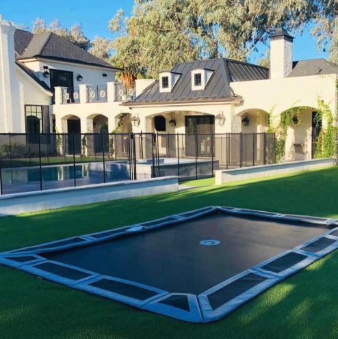 Rectangular in-ground trampoline with gray edge padding set into artificial turf in front of a large white house and a black pool fence
