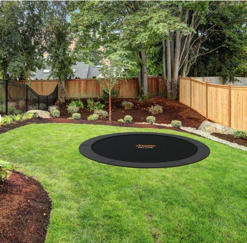 Round in-ground trampoline with an Avyna Pro-Line label on the frame, set in a curved lawn bordered by mulch beds, shrubs, and a wood privacy fence