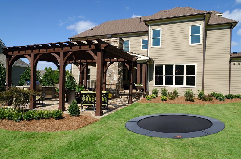 Round in-ground trampoline level with a backyard lawn next to a wooden pergola over a patio and a tan two-story house