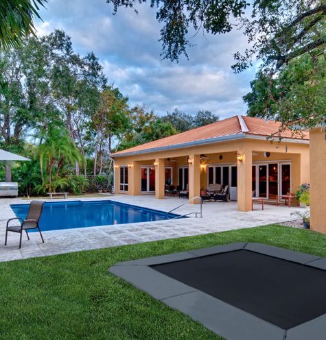 Square in-ground trampoline with gray edge padding in a lawn next to a pool deck and a covered patio, with palm trees behind