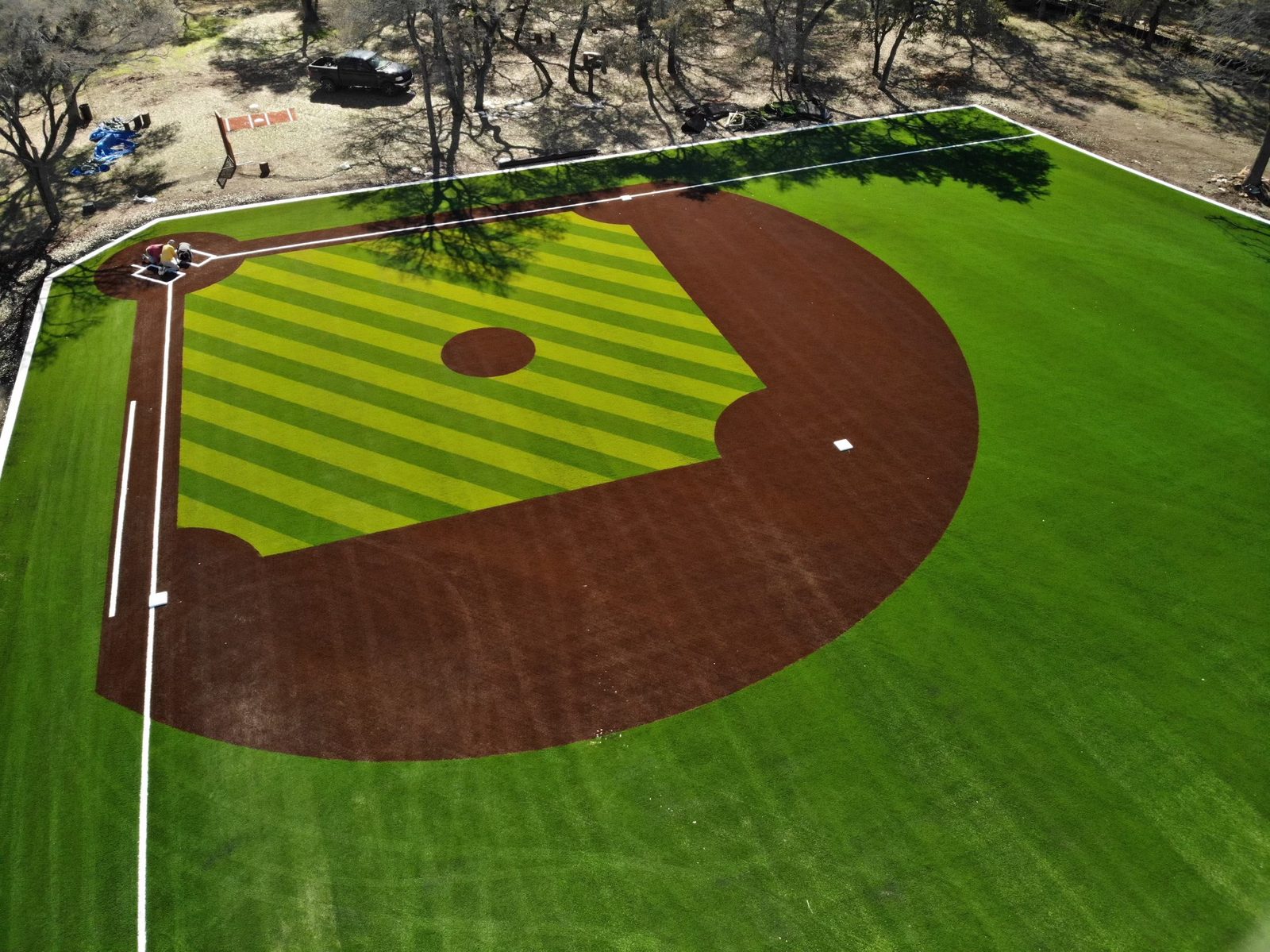 Aerial view of a striped green artificial turf baseball diamond with brown turf dirt arcs and workers near home plate