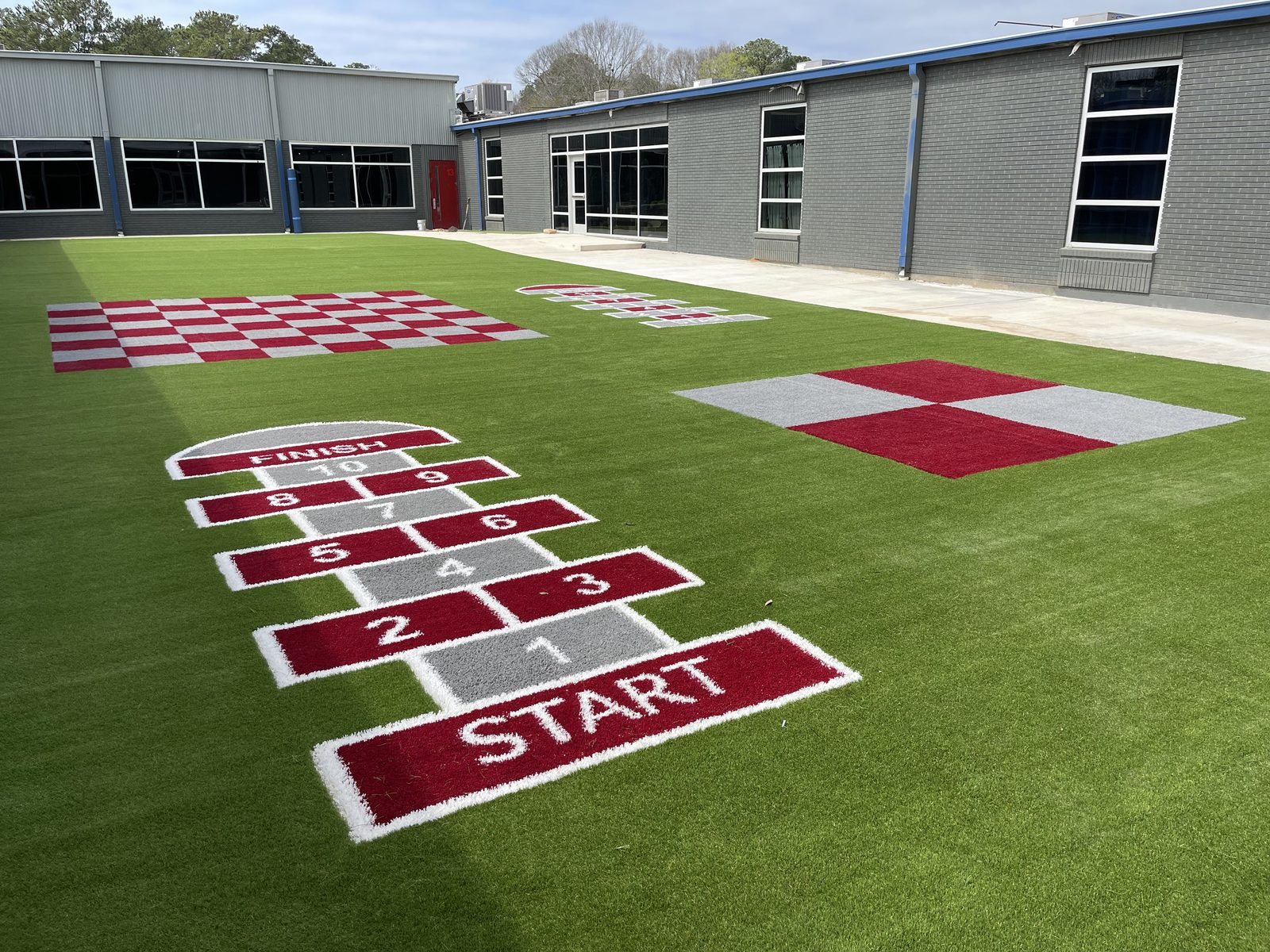 Outdoor courtyard with hopscotch, checkerboard, and four-square patterns inlaid in artificial turf beside modern buildings
