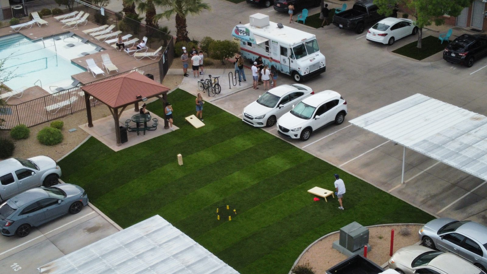 Aerial view of a striped artificial turf amenity lawn with cornhole games, a gazebo, and a food truck beside an apartment pool and parking lot
