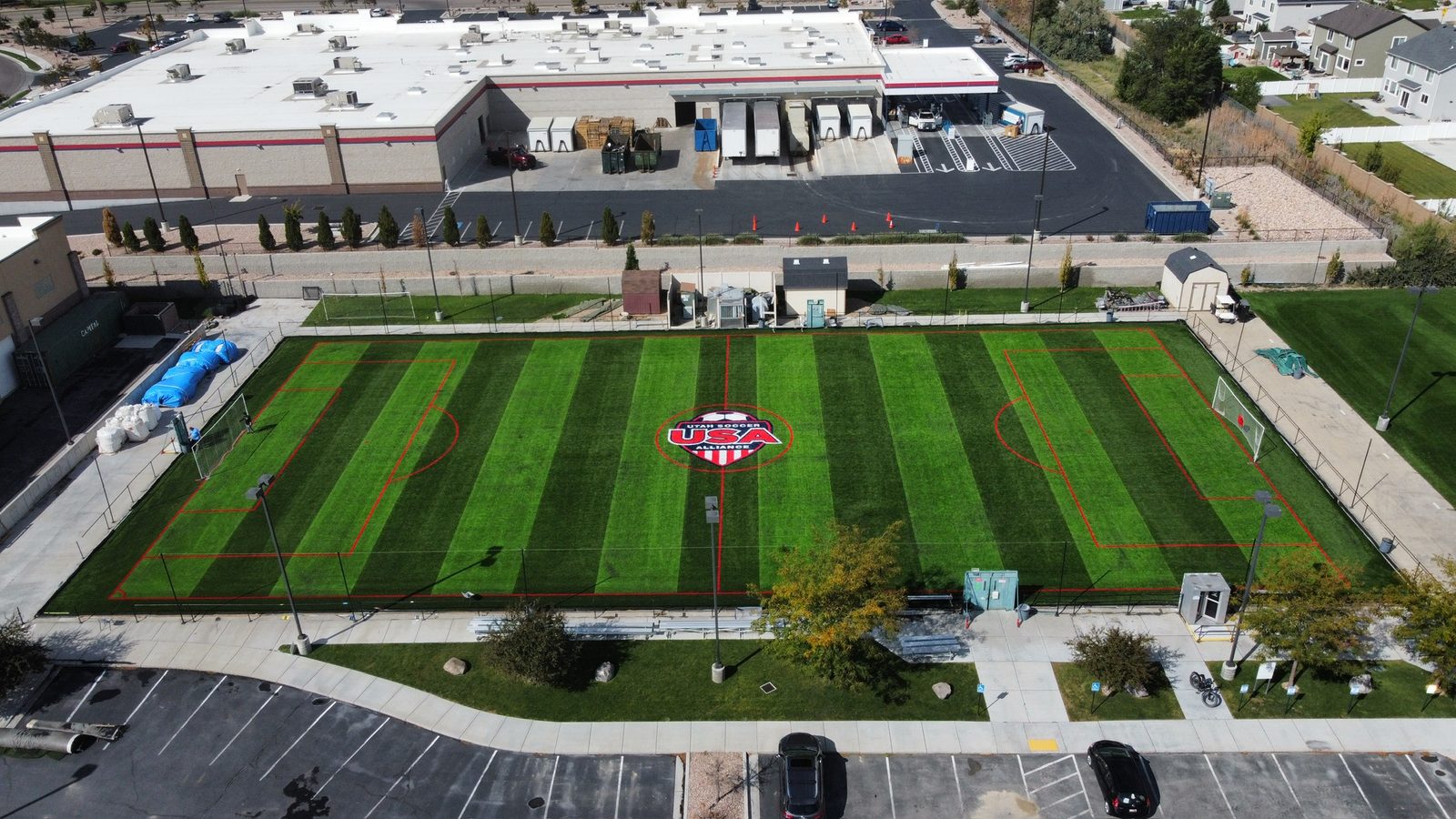 Aerial view of a striped artificial turf soccer field with red field lines and a Utah Soccer Alliance logo at center, next to a commercial building and parking lot